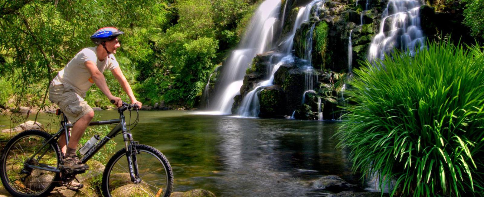 A photograph of a chap stopped along-site a waterfall on a in the bike Karangahake Gorge.