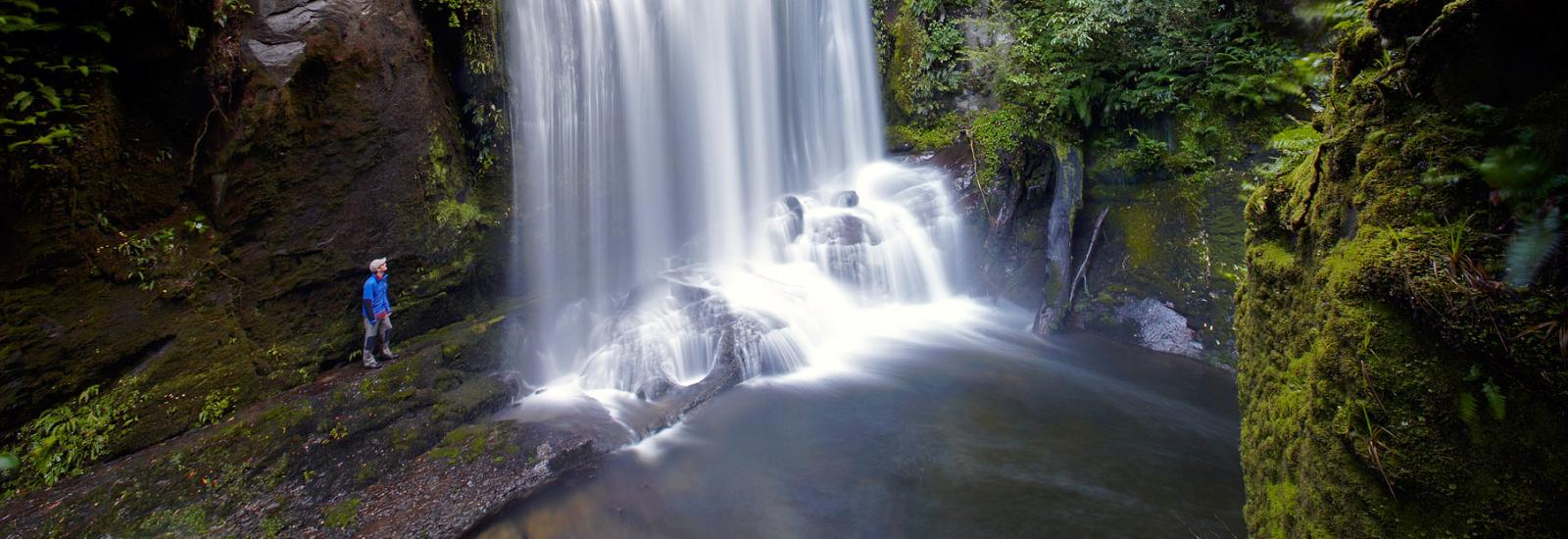 Standing next to a massive waterfall a tramper enjoys the fruits of the Lake Waikaremoana Guided Walk 
